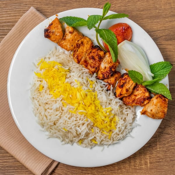 Chicken shish kebab with white and yellow rice, roasted tomato, onion, and mint on a white plate.