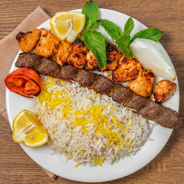 A plate of chicken and beef kebabs, rice, grilled tomato, lemon, mint, and onion.