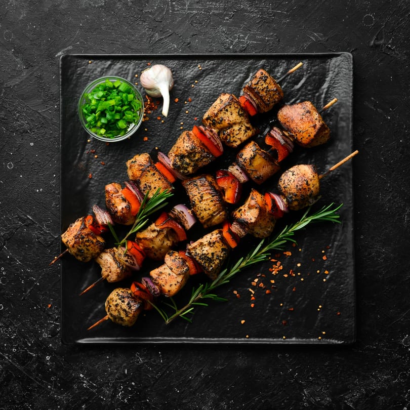 Three chicken and vegetable skewers with rosemary and spices on a black plate.